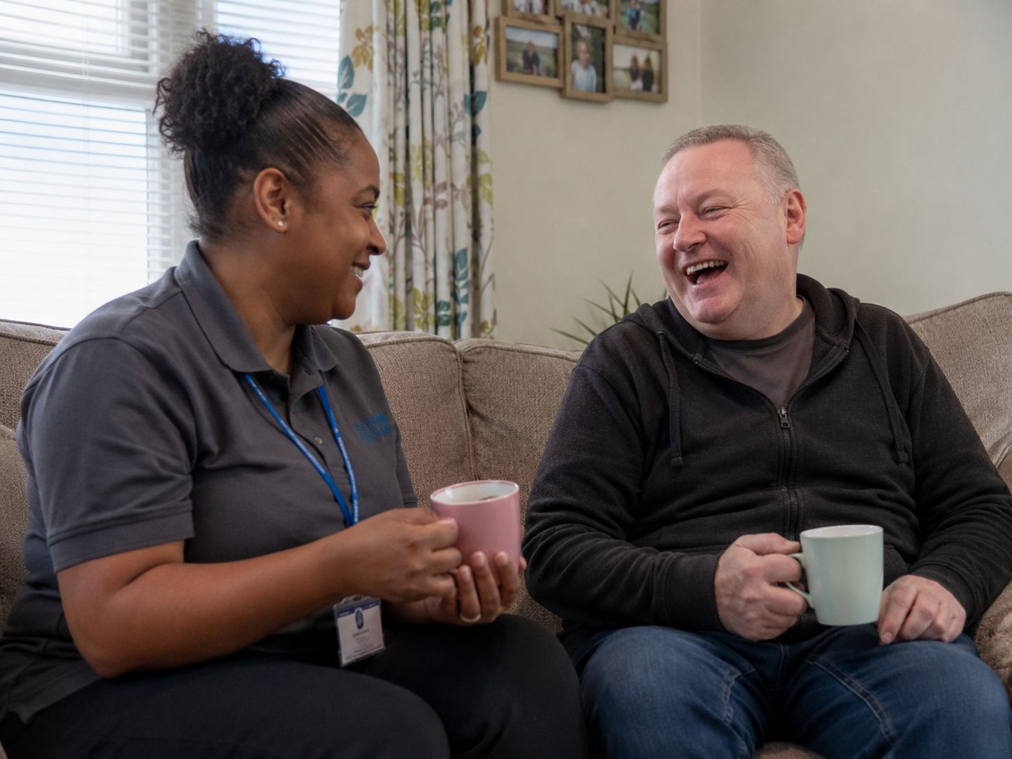 A support worker and a man sitting on a sofa at home, laughing over cups of tea